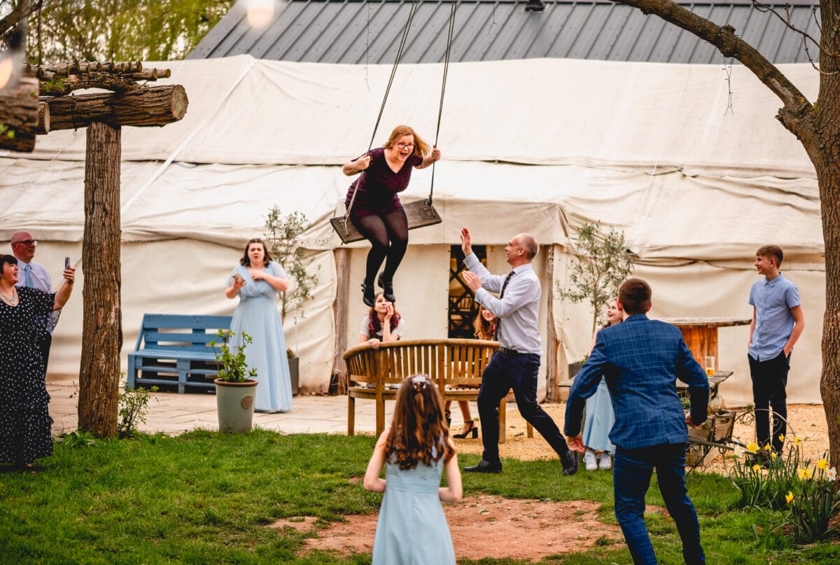Wedding guests swing on a rope swing attached to a tree at Bennetts Willow Barn, woman screams as guests laugh