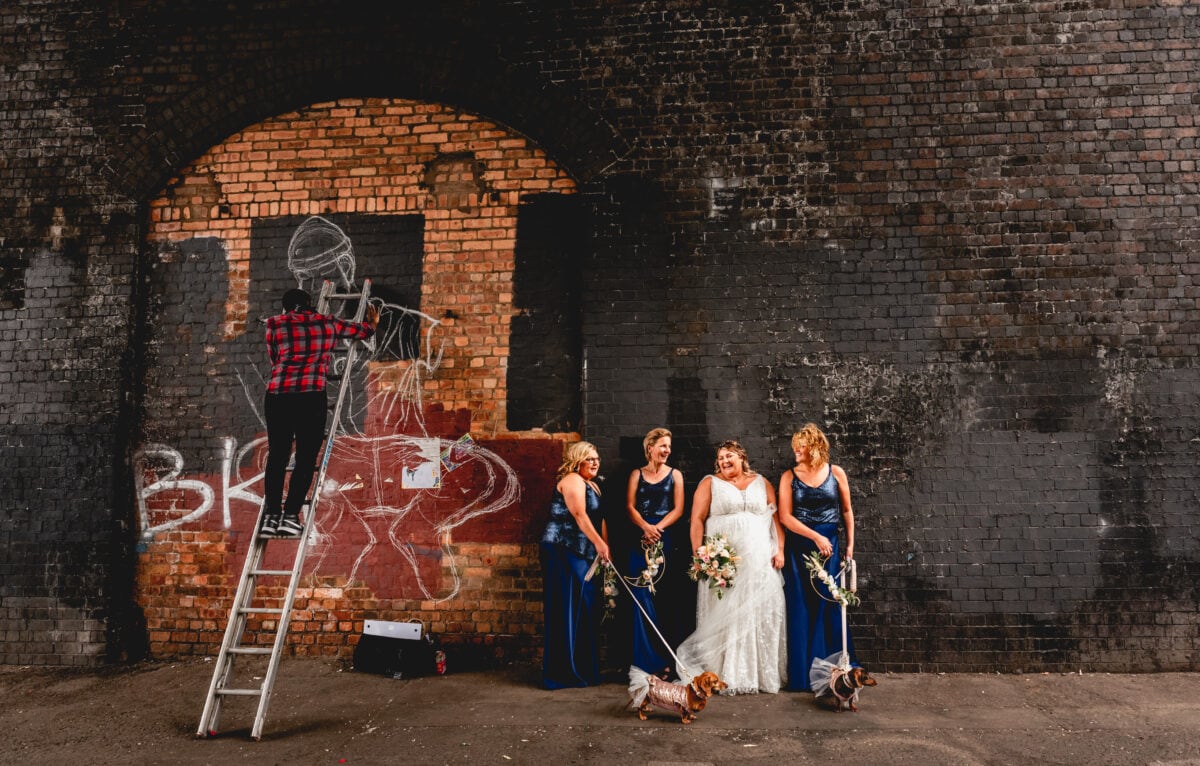 Bride and three bridesmaids in blue sequinned tops and wide leg trousers stand against a black brick wall with two pugs while a graffiti artist on a ladder paints the wall behind them