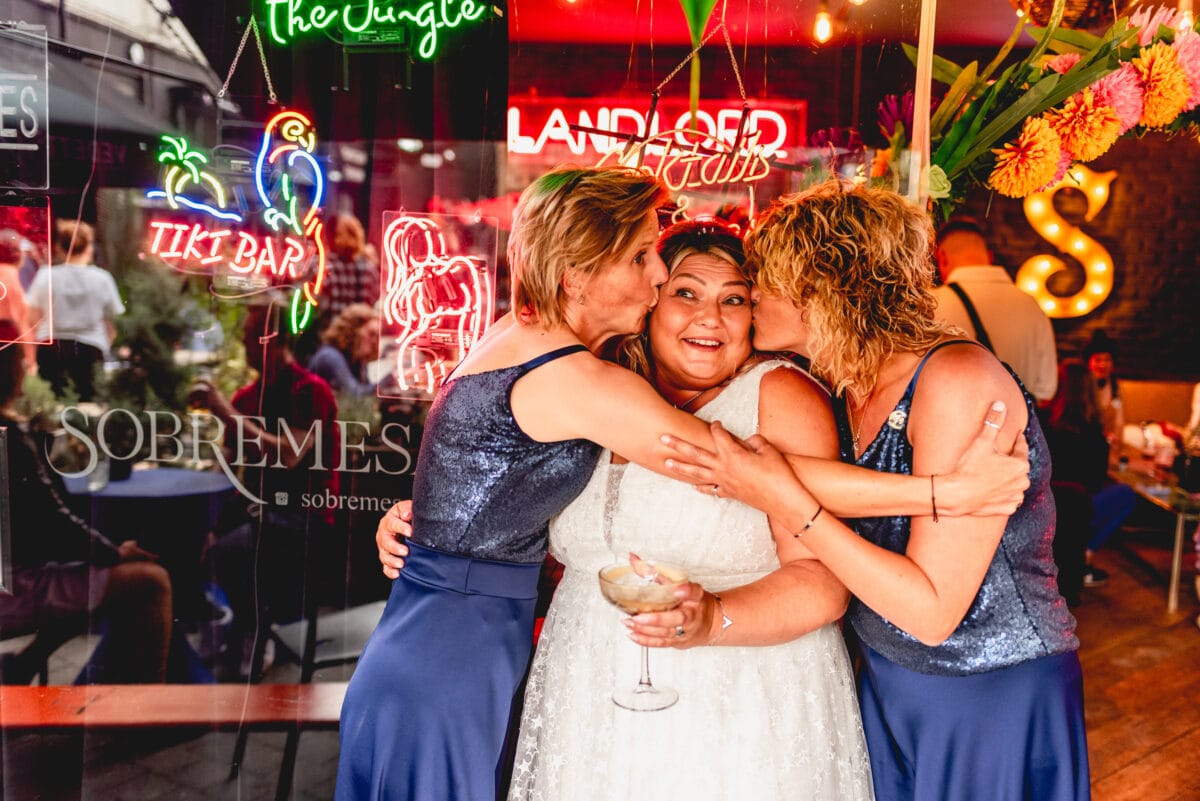 Bride is kissed on both cheeks by two bridesmaids wearing navy blue outside the Sobremesa Bar in Digbeth with neon lights in the window