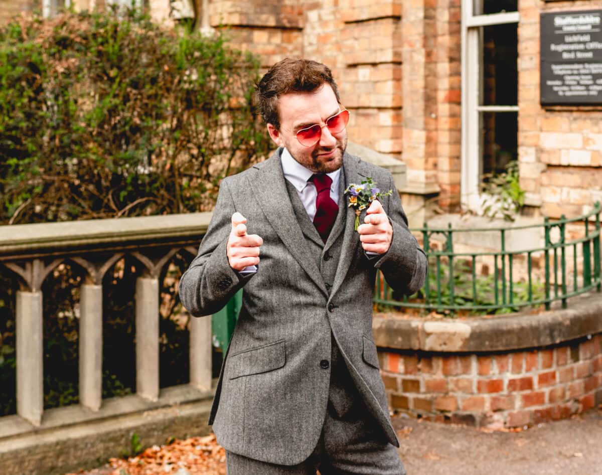 Groom in rose coloured heart shaped glasses fires finger guns at the camera