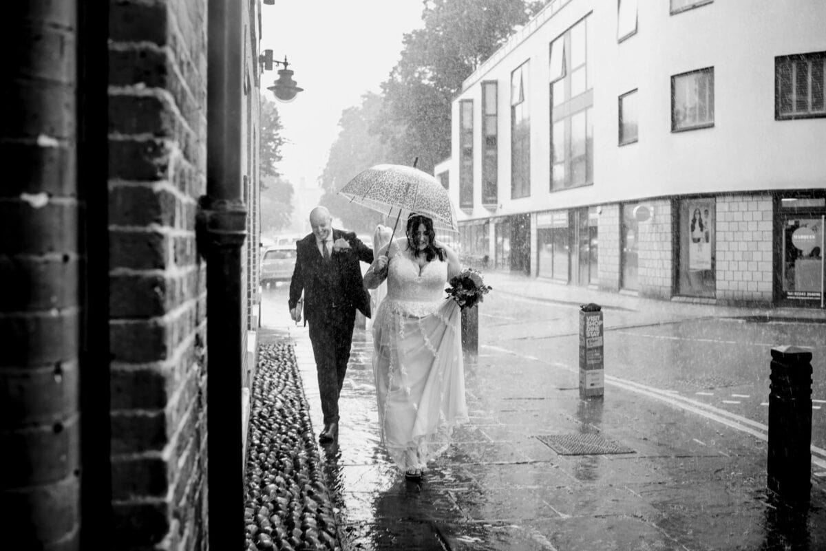 Black and white photo of bride and groom running through the rain under an umbrella on their wedding day on the streets of Lichfield City.