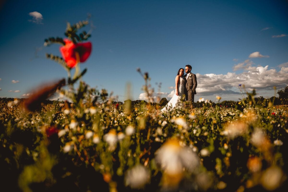 Couple stand in poppy field at Mythe Barn Wedding venue during the portrait session on their summer wedding evening