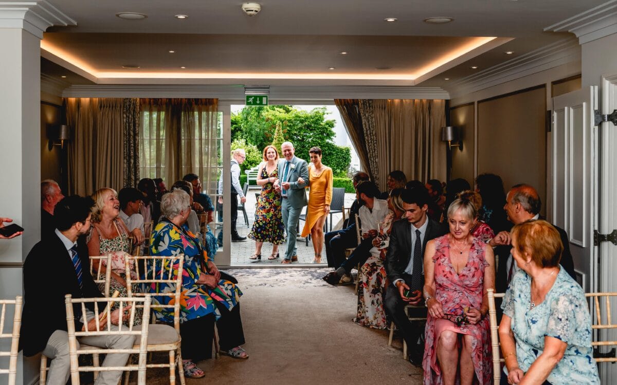 Groom enters the wedding ceremony room with his two daughters on each arm while seated guests cheer them on.