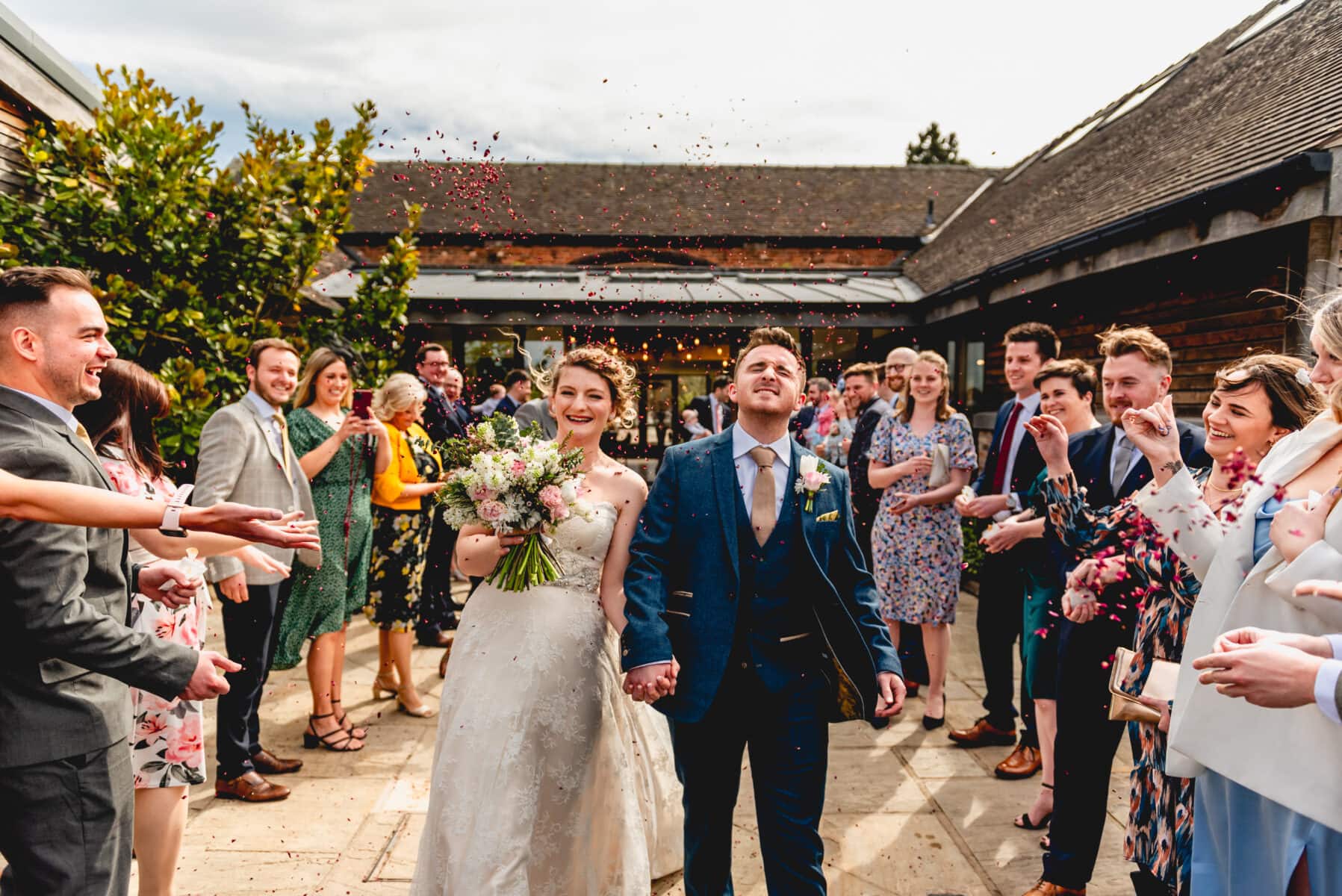 Couple pull faces as they are pelted with confetti walking through confetti tunnel at a barn venue