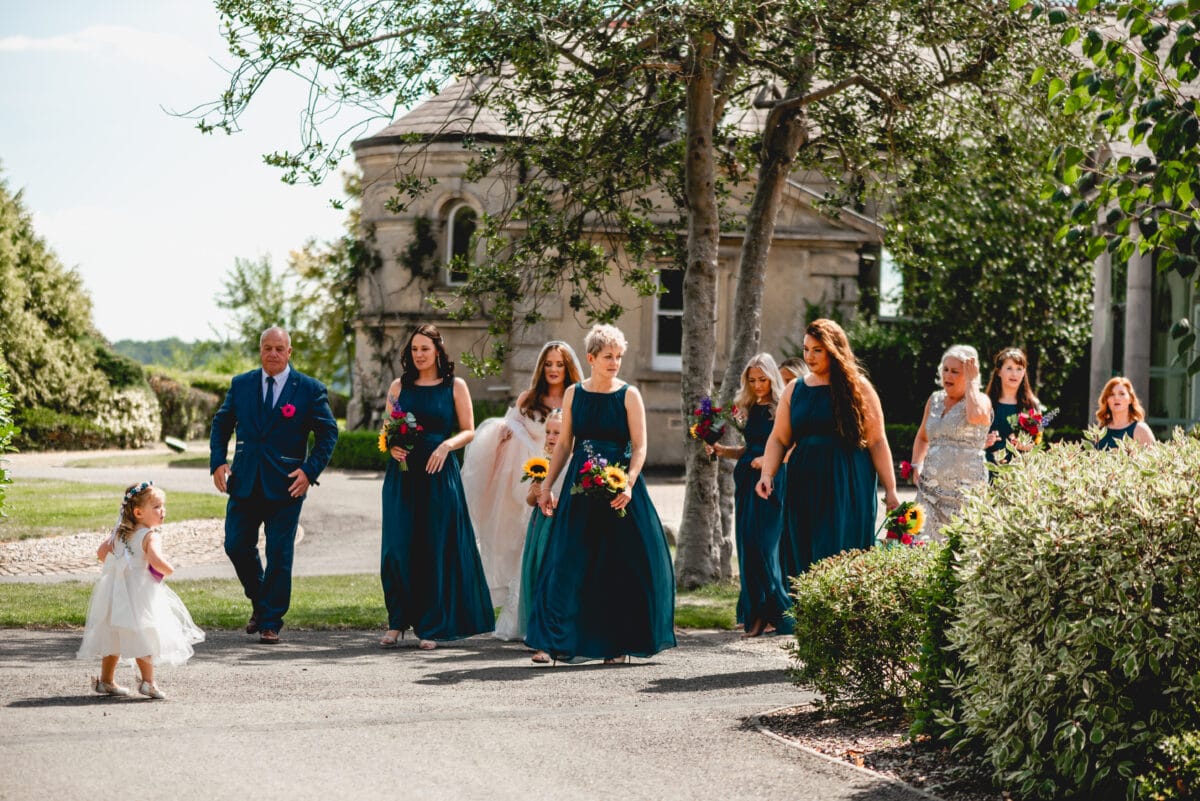 Bridesmaids and bride walk from bridal suite to outdoor ceremony area, bridesmaids in dark green dresses