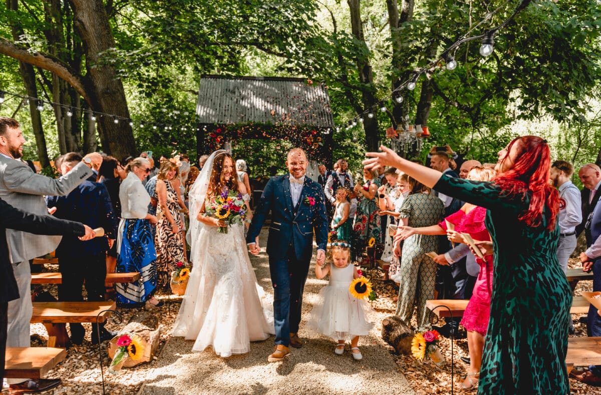 Bride and groom leave wedding and have confetti thrown at the to celebrate their outdoor ceremony, groom holds the hand of his toddler daughter who is holding a sunflower