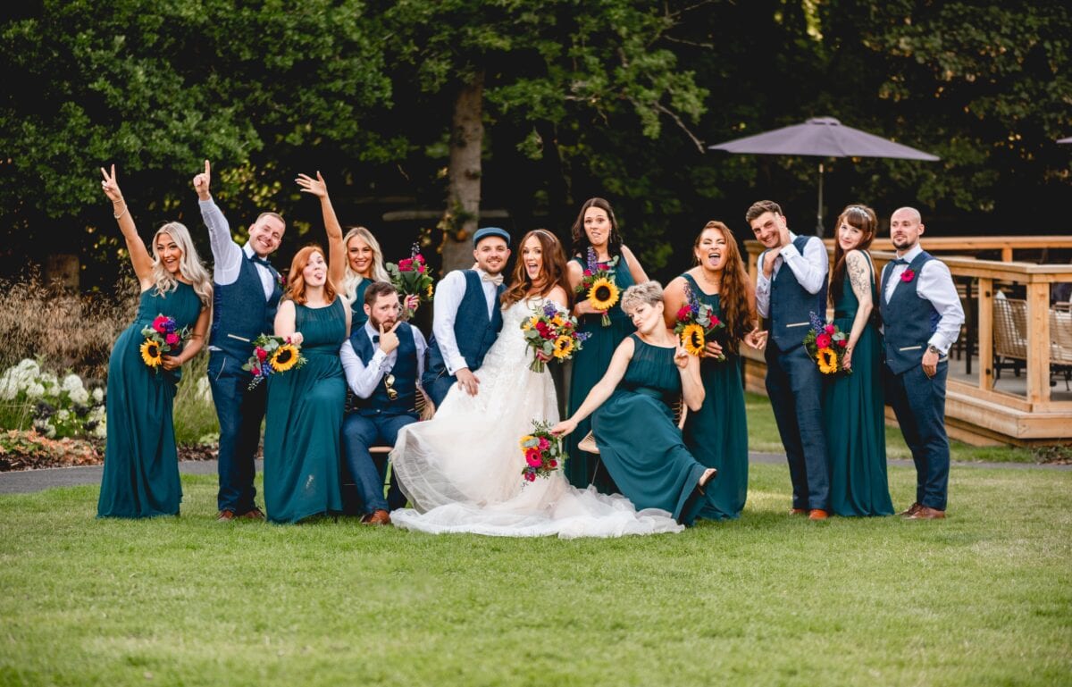 Bridal party all pose for the camera at a summer wedding, bridesmaids all wear dark green and groomsmen and ushers are all in waistcoats.