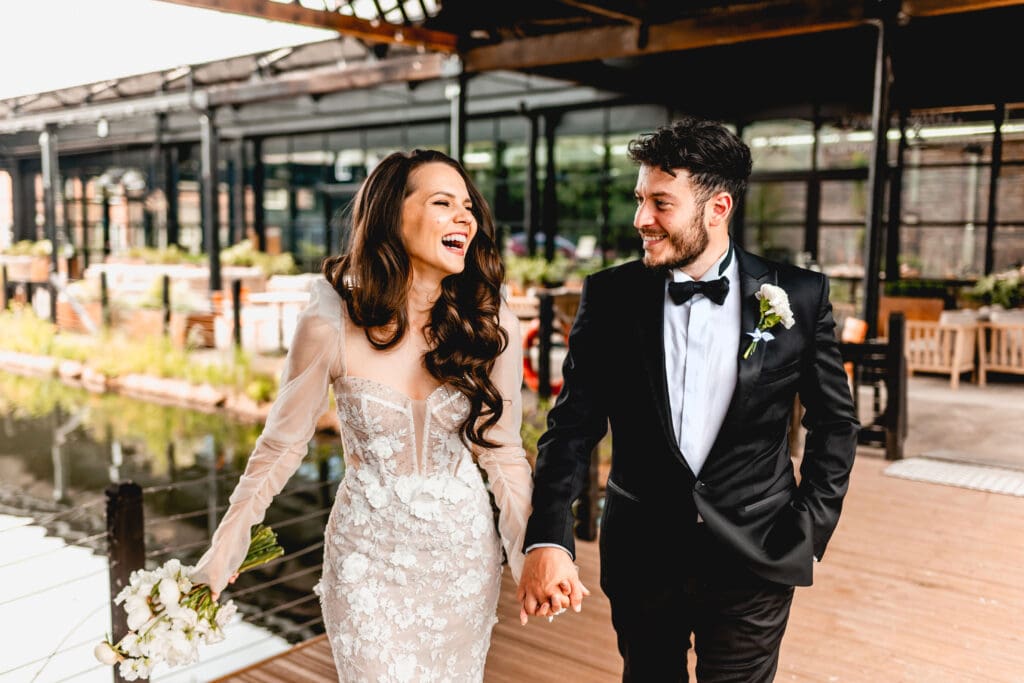 bride and groom hold hands and laugh at The Bond Company in Digbeth while walking towards camera. Bride wears Suzanne Neville dress