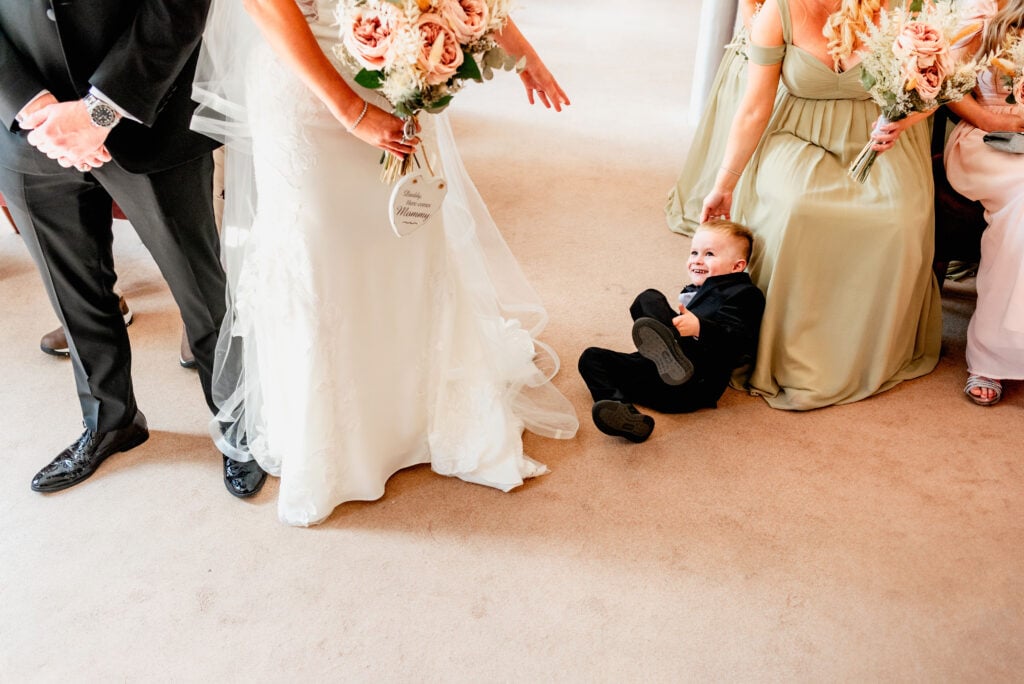 little boy lies on the floor after falling over during a wedding ceremony as his mom reaches to pick him up