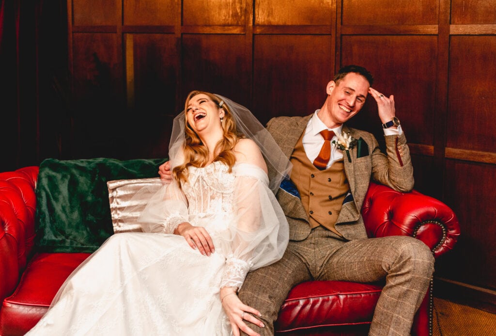 bride and groom laugh on a red leather sofa at Deckerdence wedding venue in Shustoke