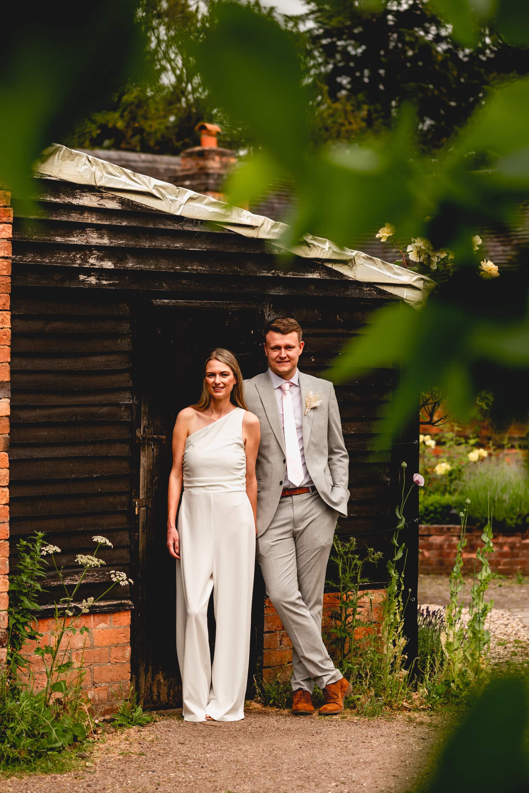 Middleton gardens bride in white jumpsuit and groom in grey suit stand against a black shed