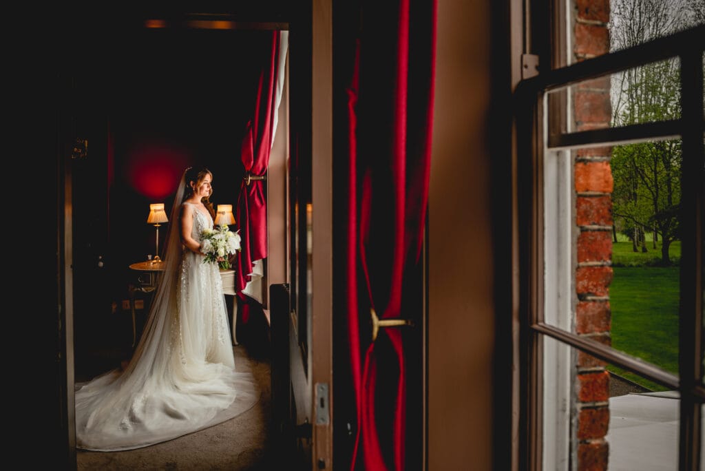 Bride looks out of a window in red velvet curtained room before her walled garden ceremony