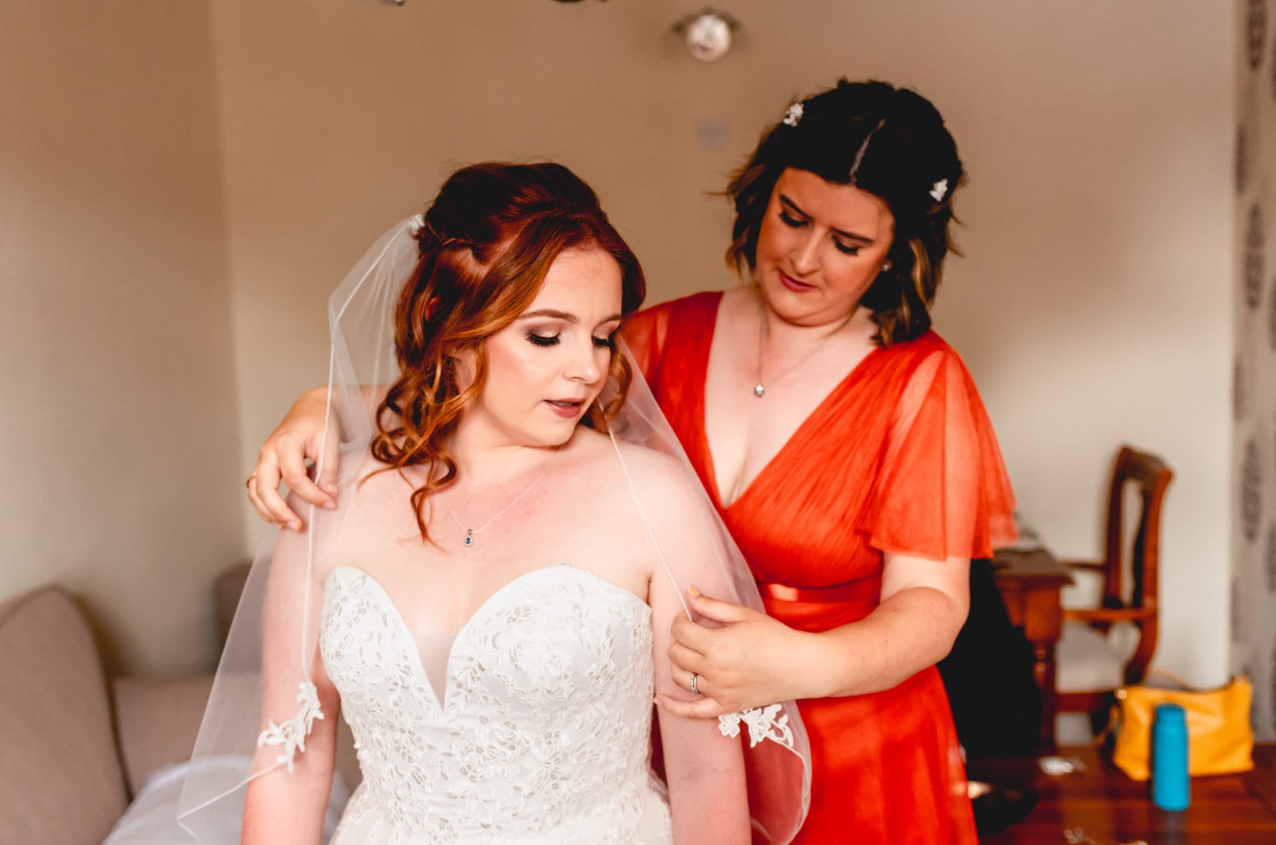 Bridesmaid in orange helps bride to adjust her veil during bridal preparation