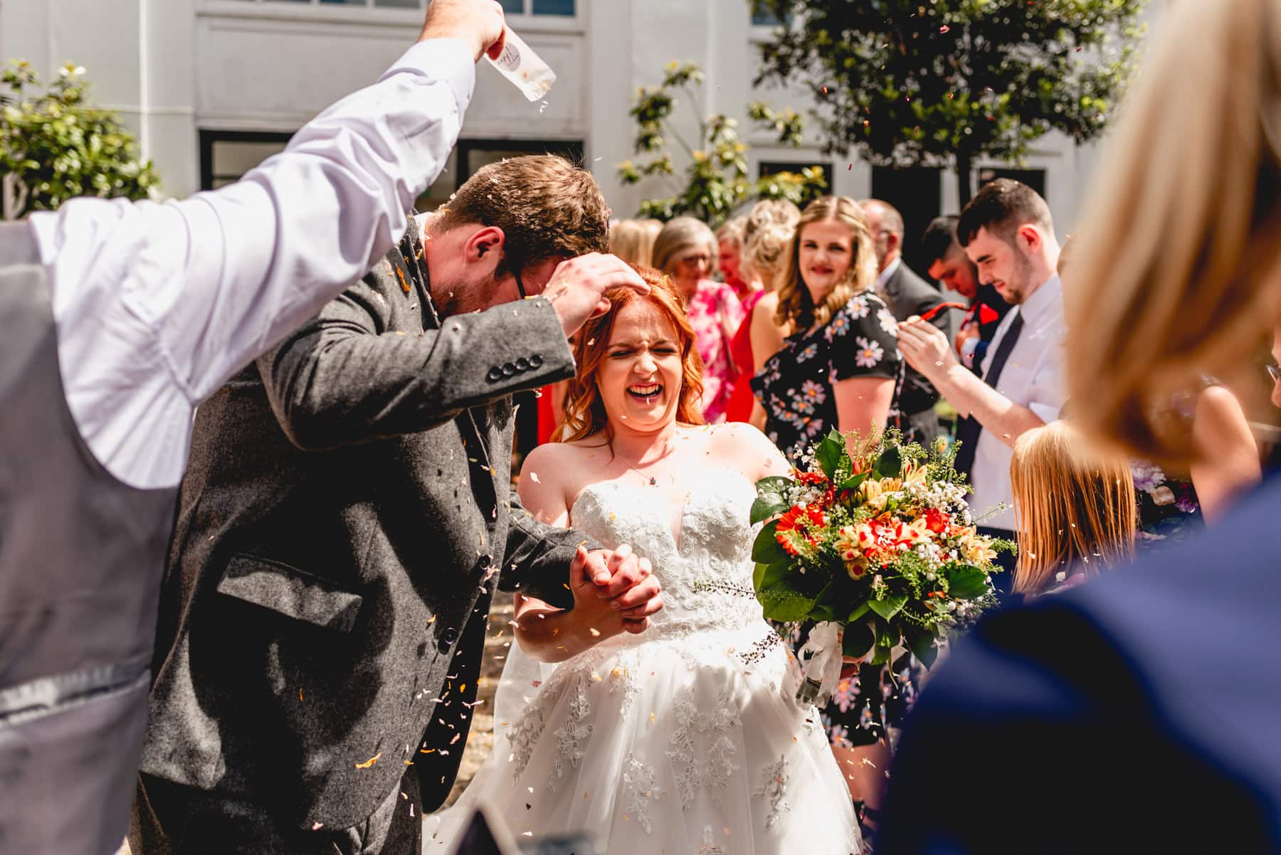 Couple have confetti petals poured over them as they laugh and hide from the armfuls of confetti