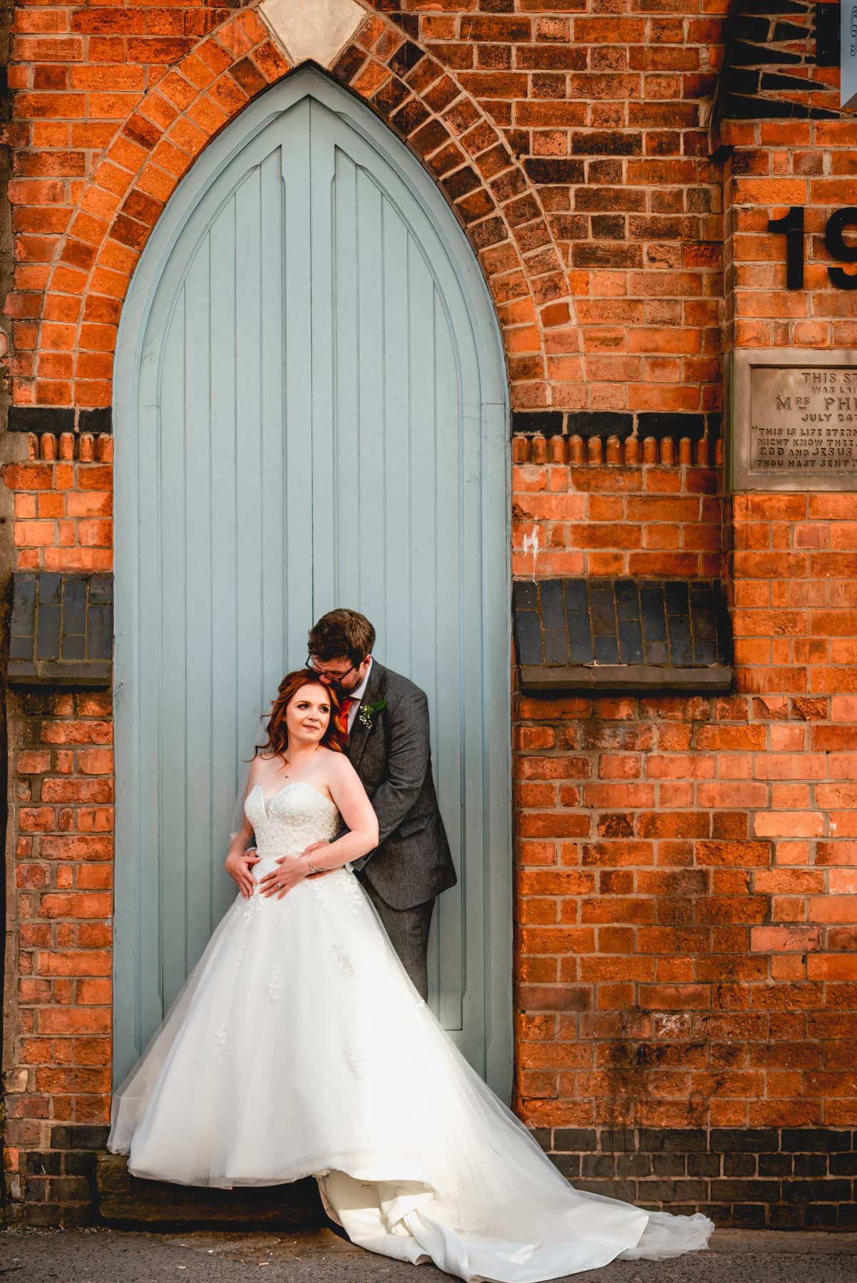 Bride in white wedding dress is held by groom against pale blue double height doors in Digbeth