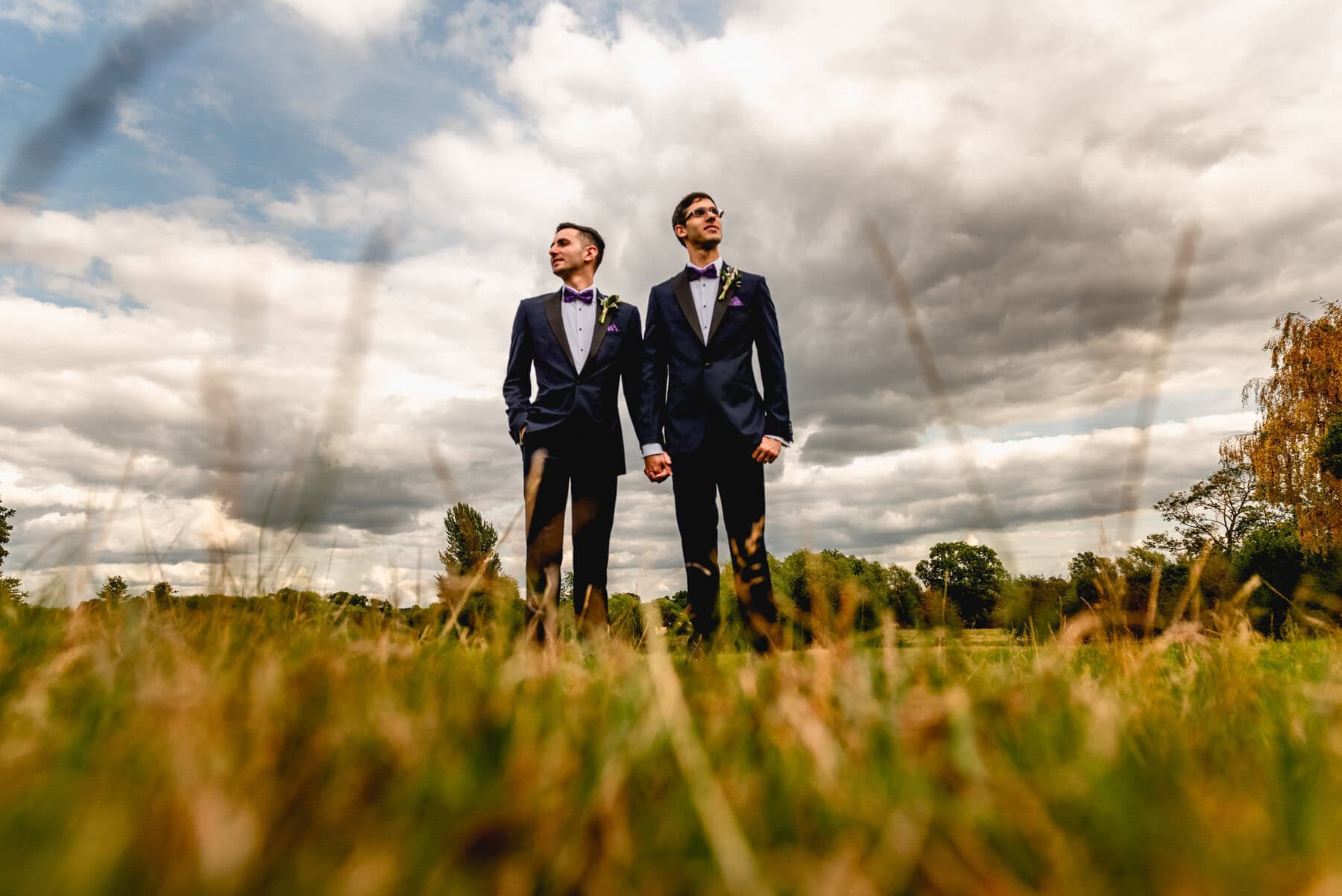 Two grooms standnin a field on their wedding day wearing blue dinner jackets and bowties at Mythe Barn Wedding Venue