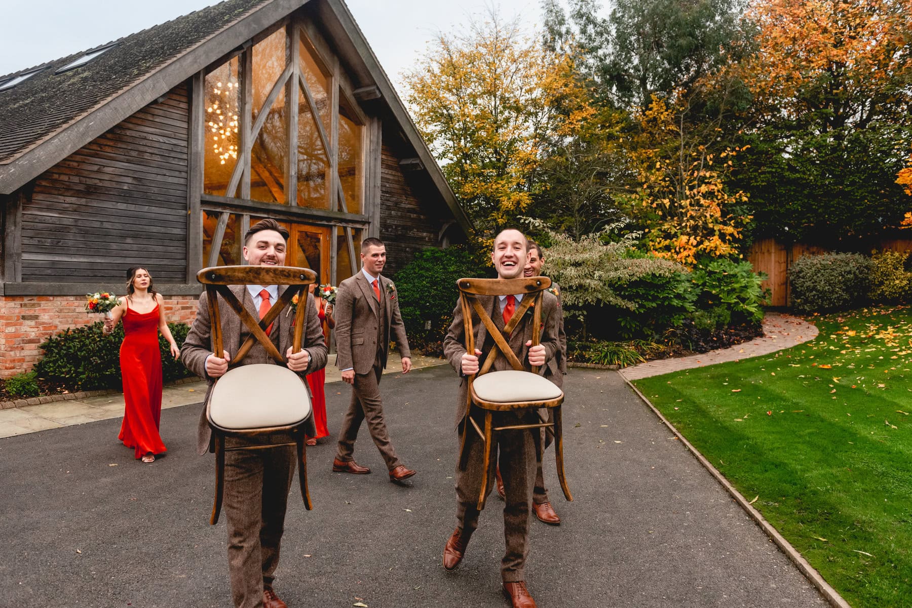 groomsmen carry chairs out of a barn wedding venue, dressed in brown tweed suits with orange ties