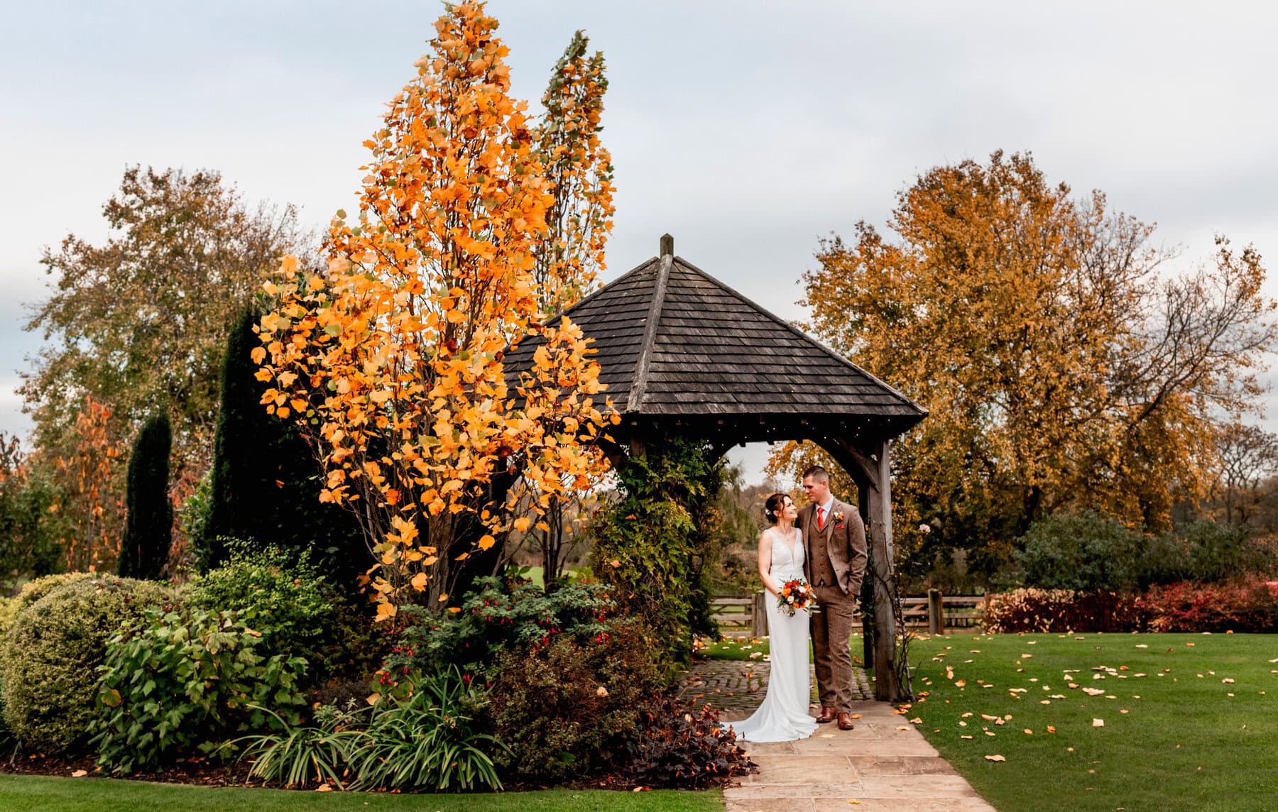 bride and groom stand under gazebo surrounded by autumn colours of reds and oranges on day of wedding