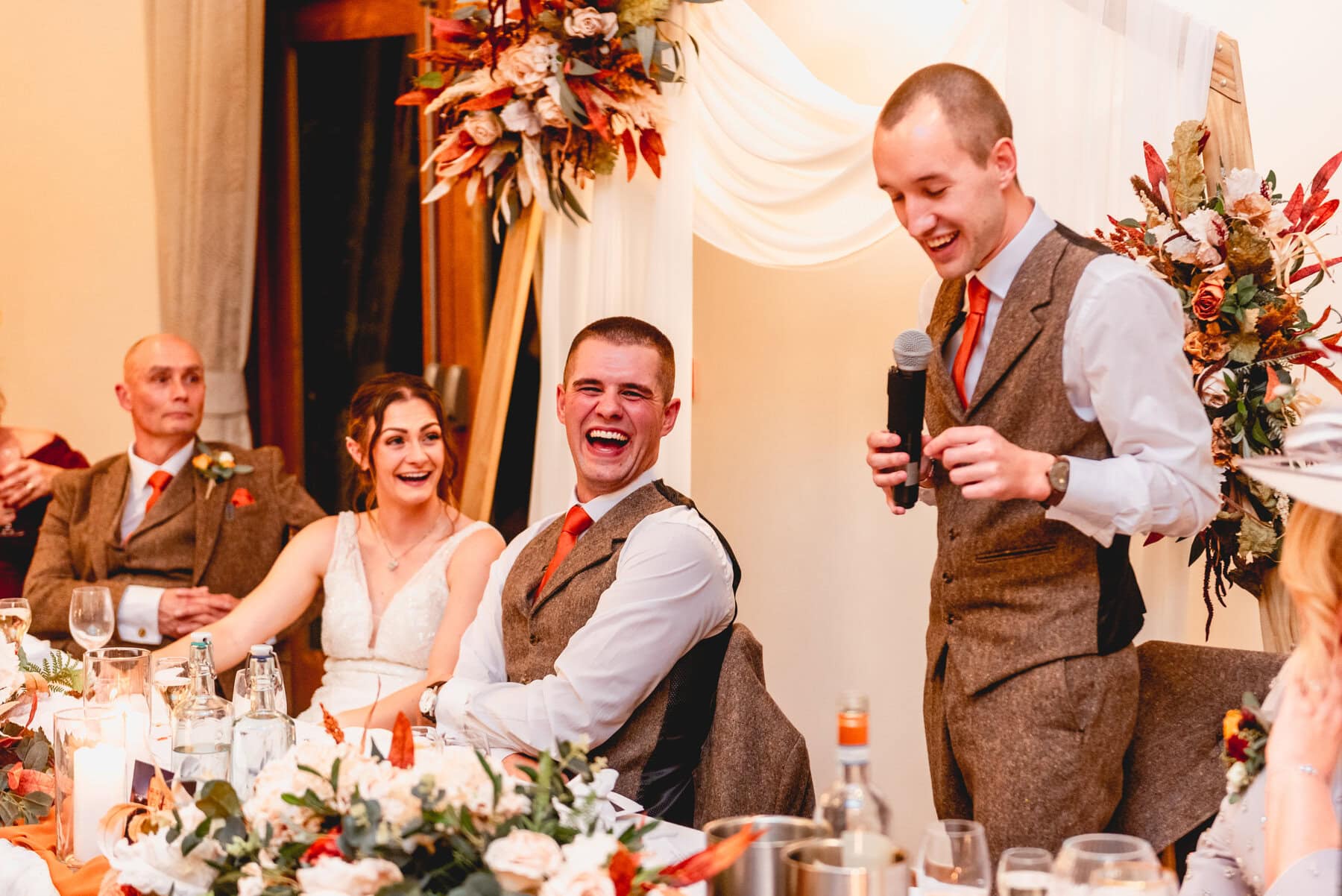 best man does speech in wedding reception while bride and groom look on laughing