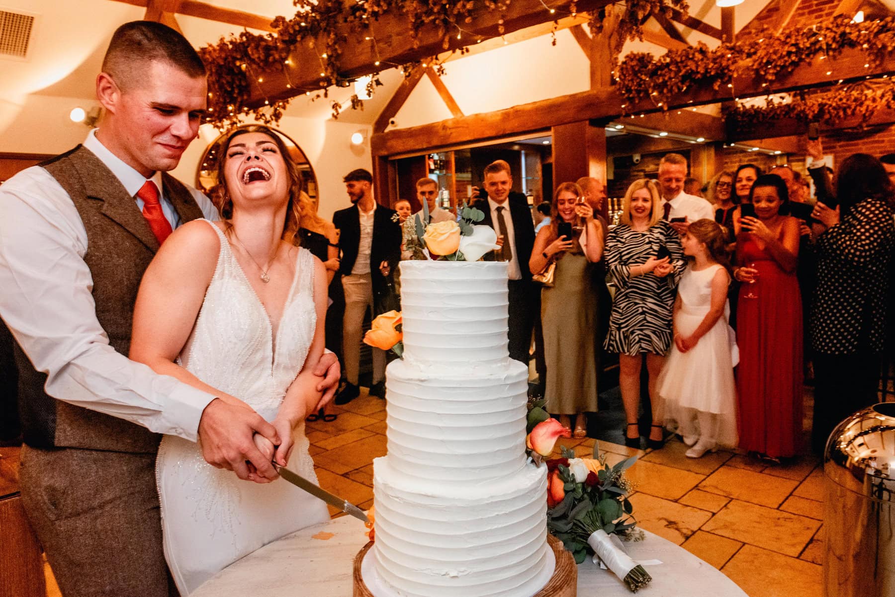 bride and groom cut cake while laughing on wedding day in barn venue with guests watching and cheering