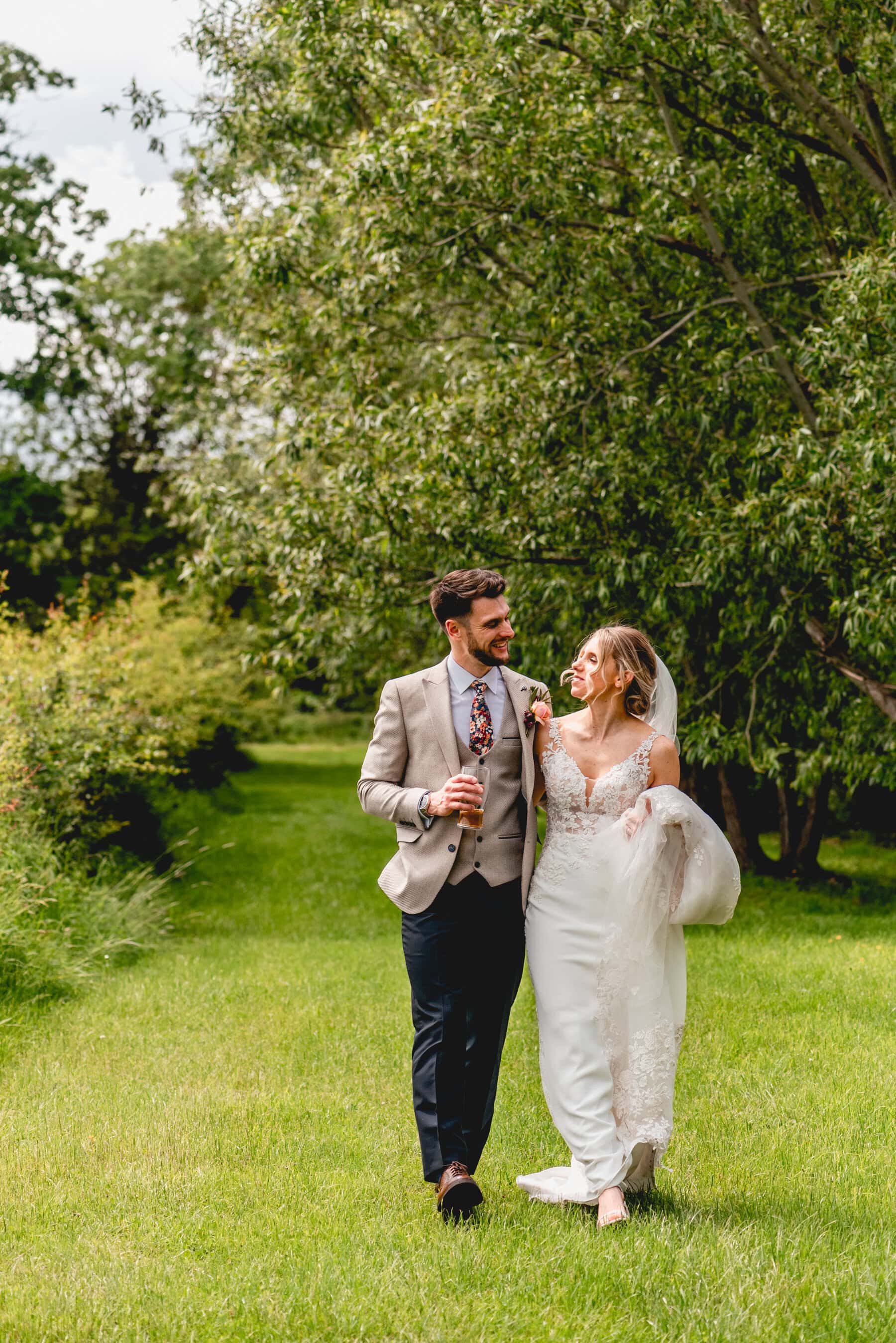 Bride and groom walk together in the field at Swallows Nest Barn as they celebrate their wedding day. Both carry a drink in their hand and bride has scooped up her wedding dress in her free hand.
