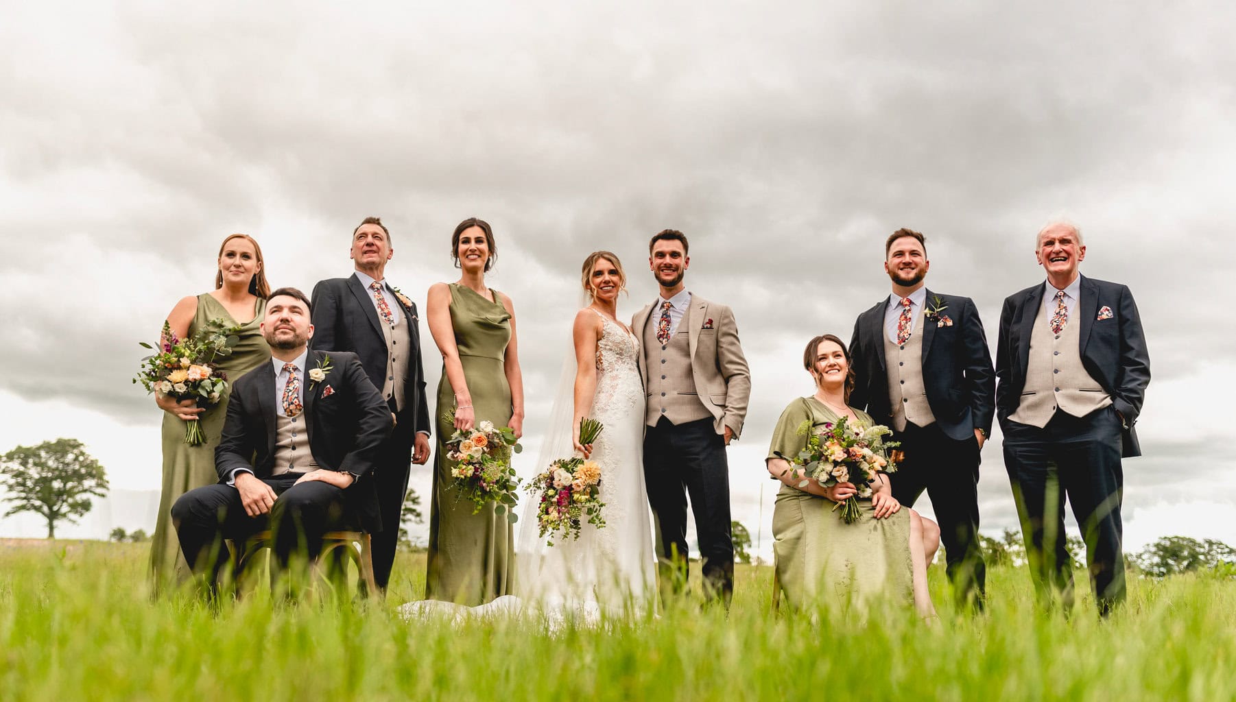 Bridal party in a field, bridesmaids wear sage green long dresses and groomsmen in suits with pink ties