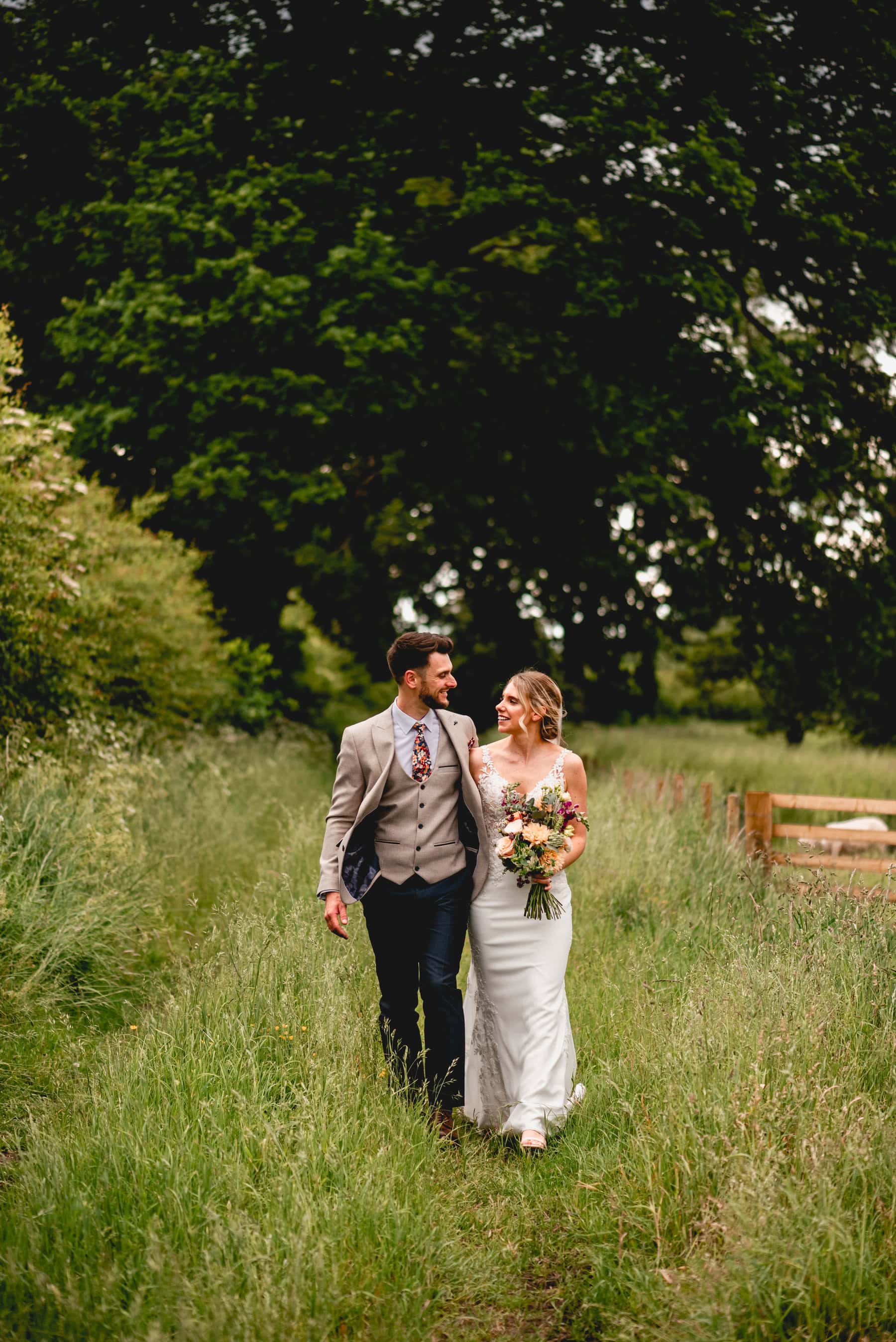 Portrait photo of bride and groom walking through the grass in a tree lined path at Swallows Nest Barn