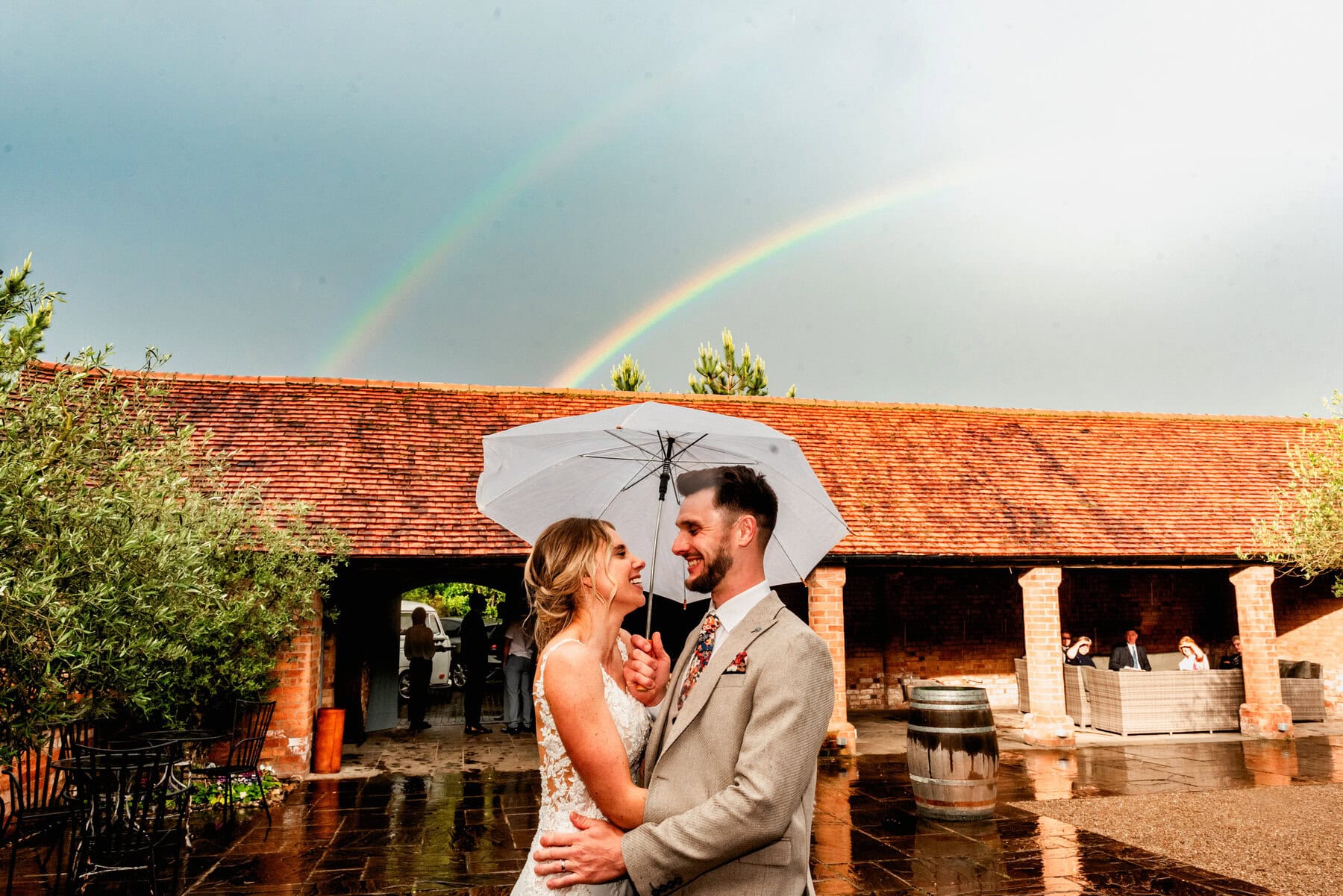 Couple at their wedding hiding under umbrella with rainbow shining in the sky behind them