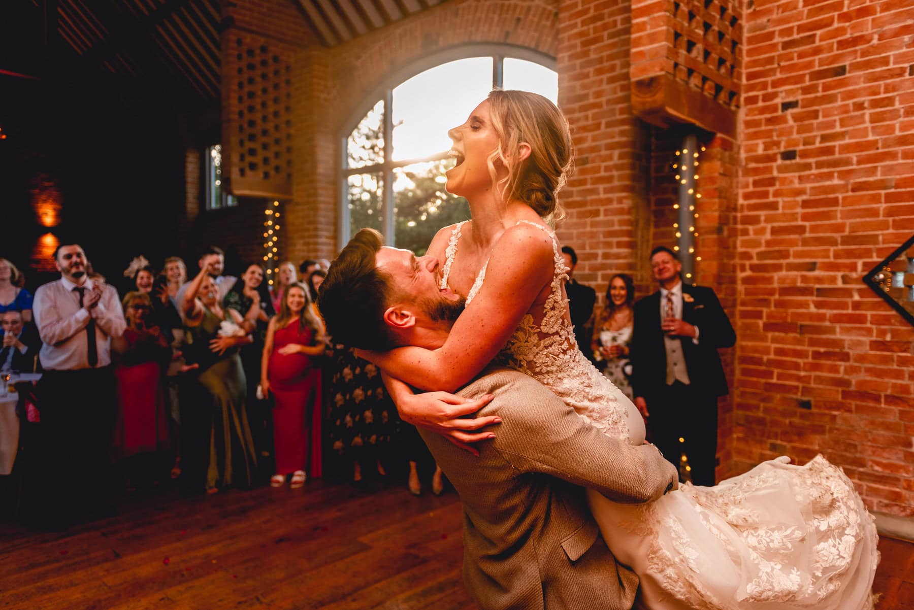 Man lifts woman into the air and twirls her around during their first dance at their wedding. The sun is setting behind the couple coating them in a golden glow