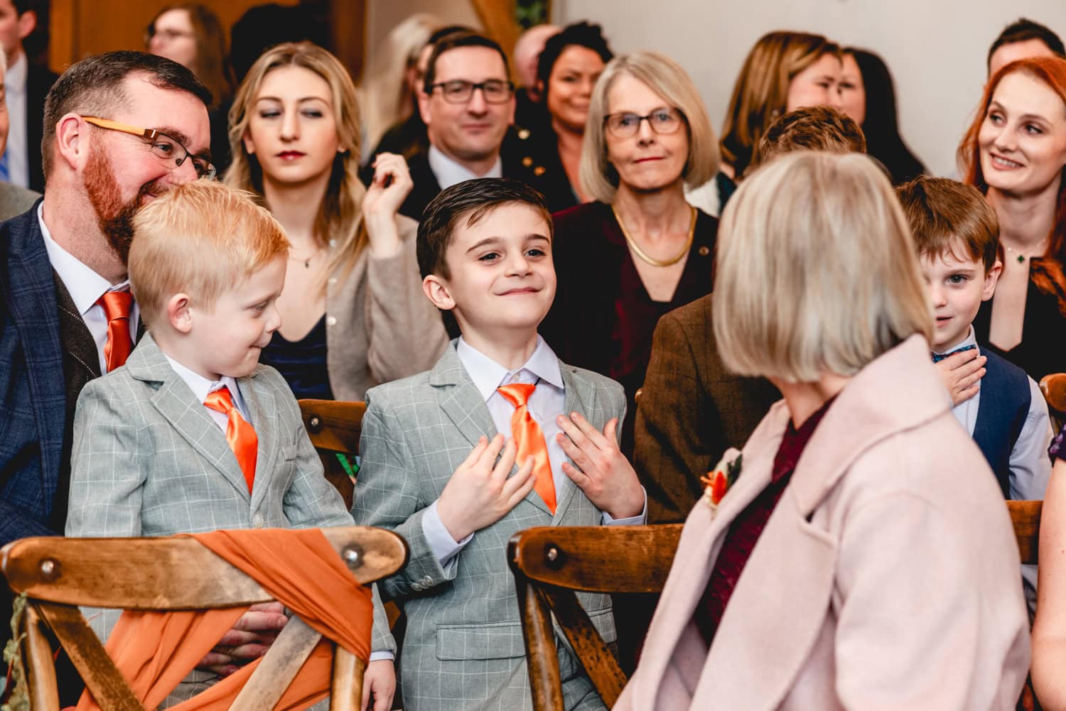 Young boy in wedding suit shows off his orange tie and grey suit to another wedding guest. 2025 - A Weird and Wonderful Year of Weddings